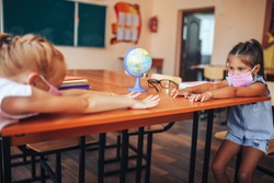 Two schoolgirls in medical masks are sitting at a school desk, opposite each other, group session, back to school, teaching children, social distance during epidemic.