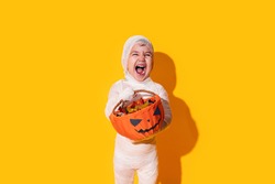 Child in mummy costume holding basket of chocolates in front of yellow background.