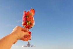 Strawberries in a glass of wine against a blue sky with a copy of space for prints, decor, wallpaper, posters. A glass of strawberries in a woman's hand.