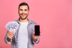 Excited man in casual t-shirt holding lots of money in dollar currencys and cell phone in hands isolated over pink wall. Phone screen for text. 
