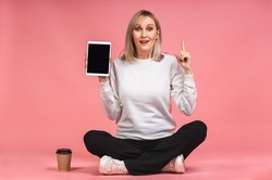 Attractive smiling beautiful young blonde woman using tablet computer, showing screen, while sitting in lotus pose on the floor isolated over pink background.