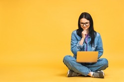 Business concept. Portrait of happy young woman in casual sitting on floor in lotus pose and holding laptop isolated over yellow background.