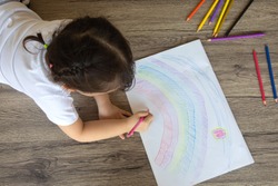 Preschooler kid is drawing colorful rainbow picture by color pencils on white paper placed on a wooden table for learning to develop creativity and enjoyment. Top view shot with copy space.