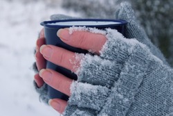 Woman holding a mug in her hands. The gloves are covered in snow. It`s freezing.Side view of frozen hands of a young woman holding a mug.