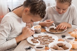 Two girls decorate halloween gingerbread cookies on plates with chocolate icing. Cooking treats for halloween celebration. Lifestyle