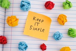 Keep it simple. A yellow paper note with the phrase Keep it simple on it with some colorful crumpled paper balls around it. Close up.