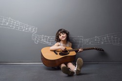 Adorable beauty plays the guitar with the light smile on her face. Big acoustic musical instrument in childish small hands. Painted with chalk stave on the neutral background.