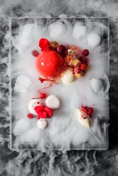 Yummy mousse cake, yogurt spheres, cherries, raspberries, a caramel decoration and fresh flowers in dry ice mist, a top-view image. Molecular food.