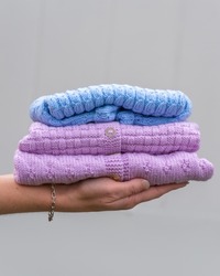 Female hand holding stacked folded baby clothes. Cozy knitted blue and pink sweaters. Handmade garment to small children. A gift to needy,  donation concept. Bright background, copy space.