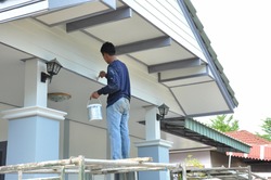 a painter one hand carry color bucket and other hand hold brush standing on scaffolding,painting house gable.worker man paint exterior house wall,people work at construction,maintenance or repair home