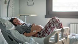 Authentic shot of an young pregnant woman in pajamas is caressing her belly few hours before to give a birth to her future baby in a hospital .