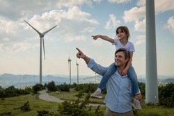 Cinematic shot of carefree young father engineer carrying his daughter on shoulders and showing windmill field. Concept of renewable energy, love for nature, family, electricity, green, future.