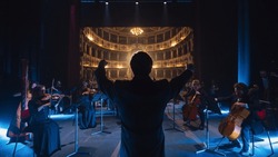 Back View Cinematic shot of Conductor Directing Symphony Orchestra with Performers Playing Violins, Cello and Trumpet on Classic Theatre with Curtain Stage During Music Concert