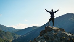 On the edge of a rocky cliff a man raises his hands to heaven as a sign of freedom or victory and in the background a fantastic landscape. Concept of: breathing, freedom, journey, life, love.