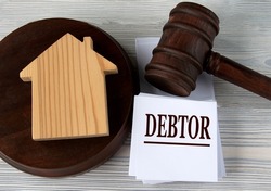 DEBTOR - word on a white sheet against the background of a judge's hammer and a wooden house.