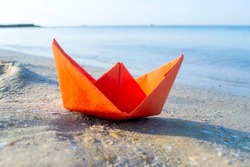 Small paper orange boat on sand near water on background of sea waves closeup. Paper boat on sandy sea beach summer sunny day. Concept adventures travelling vacation holiday rest tourism voyager dream