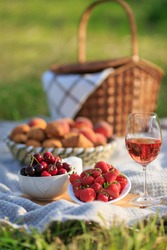 Summertime healthy picnic on nature background at sunny day. Close up Basket fruits strawberries, cherries, glass of red wine, on green grass in garden. Summer weekend outdoor. Beautiful still life