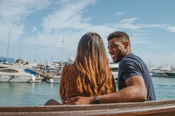Multiethnic Couple Sitting on a Bench in front of the Harbor with Boats Floating on the Water (Black Boyfriend with Watch Smiles Looking at his Girlfriend)