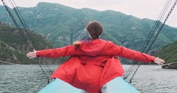 Traveler in red jacket raises arms to sides on moving boat, background of mountains landscape. Woman travel by ferry on Albania lake. Adventure, vacation concept. Explore beauty world, recreation