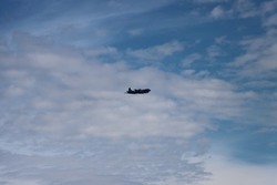 A large, grey military aircraft flying in a blue sky with light cloud cover. The sky is bright with various fluffy cloud patterns. The airplane is seen from far away. Airforce beach, Courtenay, BC.