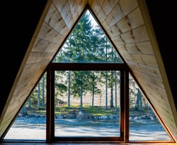 Log cabin window. Looking through the window from inside. Nature behind window glass. Window frame and curtain. Triangle. Timber construction. Wooden, log hut. Warm wooden shack. Sunny weather