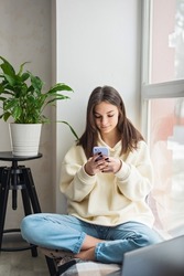 Happy teenage girl looking at smartphone, relaxing at home, enjoy using mobile online apps, playing games on mobile phone, checking messages or social media messages. Soft selective focus.