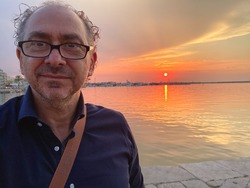 portrait of Italian man with gray curly hair and high forehead with dark blue shirt and the sea in a harbor with boats and for background the horizon with sky and red sun at sunset