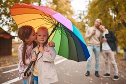 Two gay parents with their adopted daughters walking in park together. Two cute girls standing on foreground with rainbow umbrella. Happy LGBT family concept.