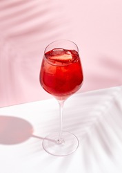 Red summer cocktail in wine glass on creative background. Shadows of palm leaf on pink wall and white floor. 
