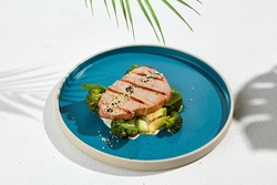 Grilled tuna steak with broccoli and zucchini in modern ceramic plate. Healthy food - roasted tuna with green vegetables. Fish dish in minimal style. Tuna fillet in blue plate with hard shadow