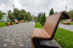 Modern wooden benches standing on the sides of the paved paths of the city park on a rainy day.
