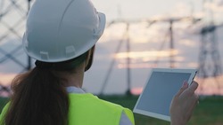 An adult woman electrician is working with a tablet on modern technologies next to a power plant, setting up energized stations through satellite control of networks, volt-installed towers, a gadget