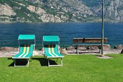 Colourful Empty Sun Beds on Grass beside Wooden Bench on Shore of Lake Garda 