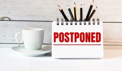The word POSTPONED is written in a white notepad near a white cup of coffee on a light background