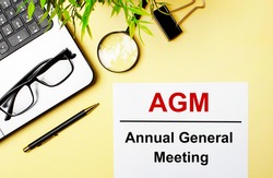 AGM Annual General Meeting is written in red on a white piece of paper on a light yellow background next to a laptop, pen, magnifying glass, glasses and a green plant.
