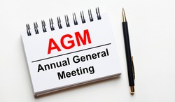 On a light background, a white notebook with are words AGM Annual General Meeting and a pen.