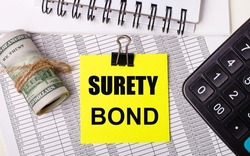 On the desktop there are reports, notepads, a calculator, a cash and a yellow sticker with the text SURETY BOND. Business concept