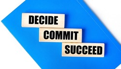 There is a blue notebook on a light gray background. Above are three wooden blocks with the words DECIDE COMMIT SUCCEED