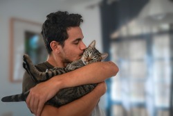 young man holds a tabby cat in his arms and kisses it