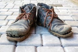 A pair of dirty boots. Old worn dark blue leather shoes with variegated brown laces. Background from gray pavers. The concept of poverty, homelessness, lack of money. Selective focus