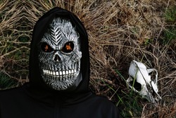 Close-up portrait of grim reaper. Man in death mask with fire flame in eyes on dark dry grass with animal skull background. Carnival costume, creepy teeth. Halloween holiday concept. Dark horror.