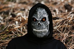 Close-up portrait of grim reaper. Man in death mask with fire flame in eyes on nature brown pine forest floor background. Carnival costume, creepy teeth. Halloween holiday concept. Dark horror.