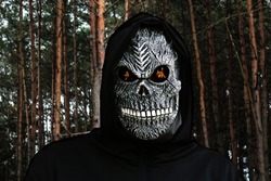 Close-up portrait of grim reaper. Man in death mask with fire flame in eyes on dark pine forest, high trunk background. Carnival costume, creepy teeth. Halloween holiday concept. Dark horror.