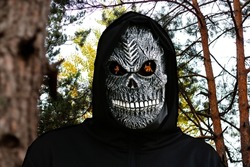 Close-up portrait of grim reaper. Man in death mask with fire flame in eyes on nature forest trunk background. Carnival costume, creepy teeth. Halloween holiday concept. Dark horror.
