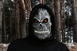 Close-up portrait of grim reaper. Man in death mask with fire flame in eyes on dark black woodland forest background. Carnival costume, creepy teeth. Halloween holiday concept. Dark horror.