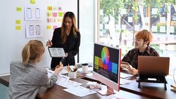 Female website designer showing user interface design on digital tablet to creative team at office presentation.