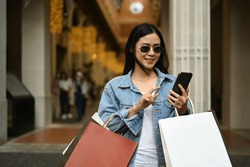 Beautiful fashionable asian woman carrying shopping bags and using smart phone outdoor front of the shopping center