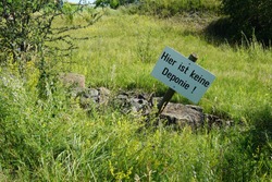 Warning sign in German language, Here is not a dumping ground, situated among stones and grass. Alarm board has rectangular shape, is made from wood painted white with black letters on wooden stick.
