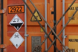 Orange container lashed with lashing bars and twist locks on deck of container ship with stickers indicated dangerous goods inside. Class 6 is toxic and infectious and class 8 corrosive substances.