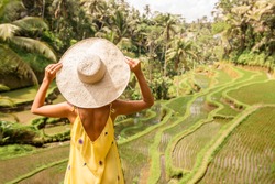 Beautiful young lady in shine through dress touch straw hat. Girl walk at typical Asian hillside with rice farming, mountain shape green cascade rice field terraces paddies. Ubud, Bali, Indonesia.